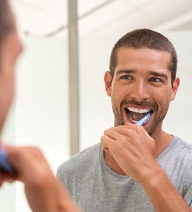 A young man wearing a gray t-shirt brushes his teeth in front of the mirror in his bathroom