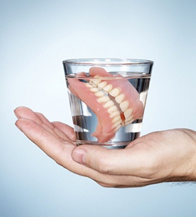 A person’s hand holding a cup full of water that contains full dentures