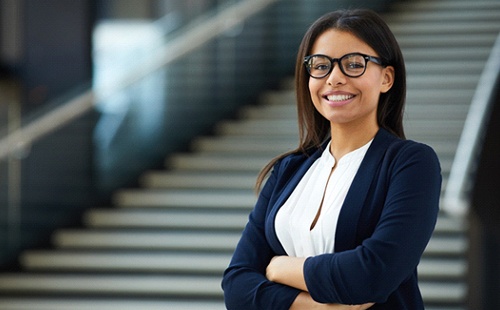 A young female wearing a navy blazer and glasses, smiling after receiving a dental implant in Delafield