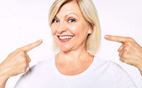 An older woman wearing a white shirt and pointing to her smile after receiving dental implants in Delafield