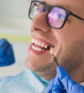 A young man wearing glasses and smiling while his dentist uses a shade guide to determine the correct color of his new dental restoration