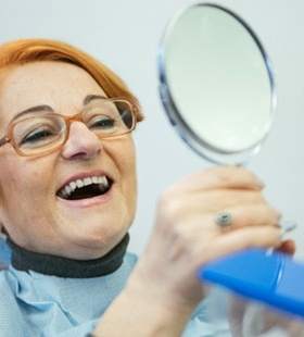 An older woman with red hair and glasses eyeing her new smile in the mirror at the dentist’s office