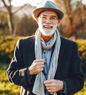 An older man wearing a coat, hat, and scarf smiling outdoors and showing off his new and improved smile