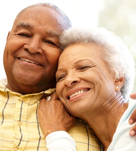 An older couple seated inside, hugging, and smiling while enjoying the benefits of dental implants
