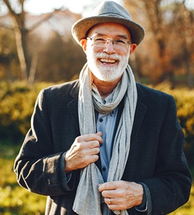 An older man outside wearing a coat, scarf, and hat while smiling and showing off his new dental implants