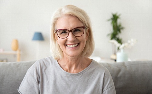 An older woman wearing a gray t-shirt and glasses, smiling after receiving a dental implant in Delafield