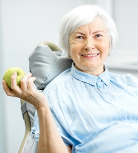 An elderly woman sitting in a dentist’s chair and smiling while holding a green apple in her hand